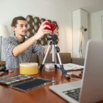 A male vlogger adjusts his camera on a tripod, preparing for a video shoot in a stylish home studio.