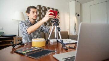 A male vlogger adjusts his camera on a tripod, preparing for a video shoot in a stylish home studio.