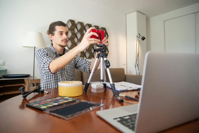 A male vlogger adjusts his camera on a tripod, preparing for a video shoot in a stylish home studio.
