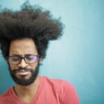 Young bearded ethnic male with creative Afro hairstyle wearing eyeglasses and pink t shirt looking down pensively thinking about trouble or question