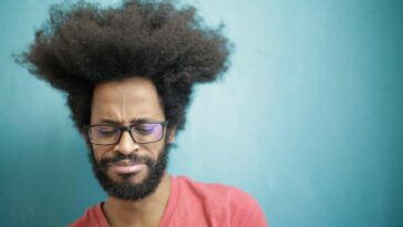 Young bearded ethnic male with creative Afro hairstyle wearing eyeglasses and pink t shirt looking down pensively thinking about trouble or question