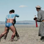 Two men in traditional attire wrestling outdoors with a referee nearby.
