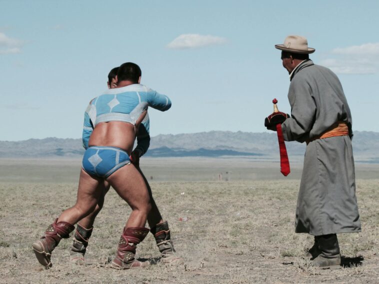 Two men in traditional attire wrestling outdoors with a referee nearby.
