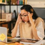A woman with glasses shows stress while working at a laptop in an office environment.