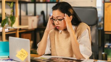 A woman with glasses shows stress while working at a laptop in an office environment.
