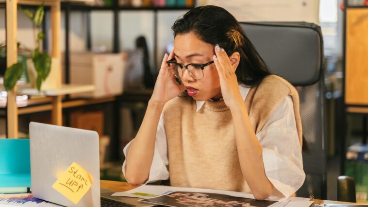 A woman with glasses shows stress while working at a laptop in an office environment.