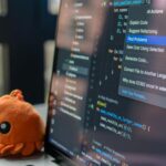 A laptop screen showing a code editor with a cute orange crab plush toy beside it.