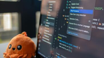 A laptop screen showing a code editor with a cute orange crab plush toy beside it.
