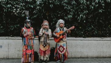 Three musicians in traditional attire performing with guitar and flute outdoors.
