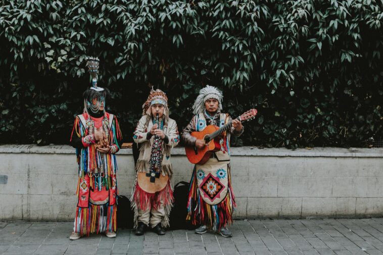 Three musicians in traditional attire performing with guitar and flute outdoors.