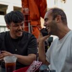 Two men smiling and chatting over drinks at an outdoor café.