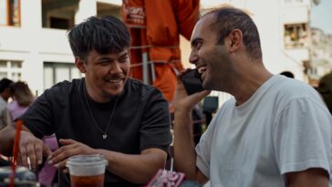 Two men smiling and chatting over drinks at an outdoor café.