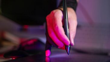 A digital artist's hand working on a graphics tablet with a stylus, in a creative studio setting.