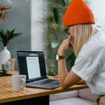 Young woman in orange beanie working on a laptop with a content plan in a cozy home office setting.