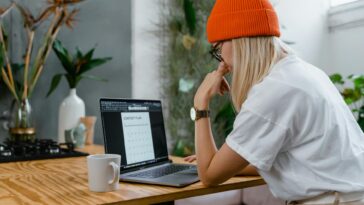 Young woman in orange beanie working on a laptop with a content plan in a cozy home office setting.