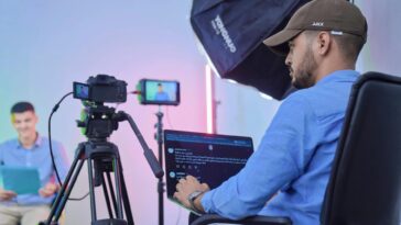 Man operates camera and laptop during a studio video shoot with a host and camera crew.