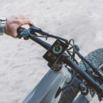 A modern electric bicycle handlebar with a touch screen display, featuring riding statistics on a sandy outdoor surface.