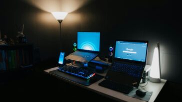 A sleek gaming desk with two laptops, RGB keyboard, and ambient lighting for a modern tech setup.