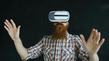 Bearded man in plaid shirt using virtual reality headset indoors, immersed in VR experience.