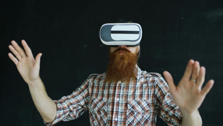 Bearded man in plaid shirt using virtual reality headset indoors, immersed in VR experience.