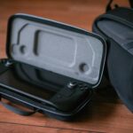 A close-up of a portable gaming device in a protective case next to a bag on a wooden floor.