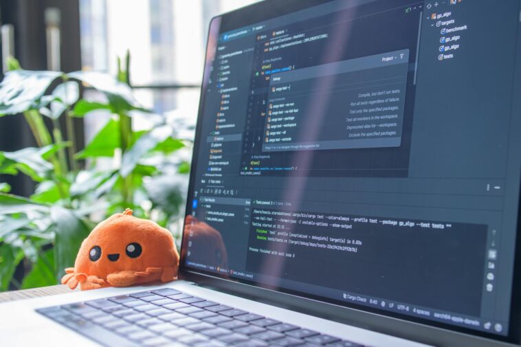 Open laptop with code editor displaying, next to an orange plush toy and green plants.