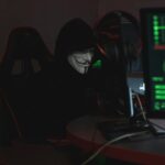 An anonymous hacker wearing a Guy Fawkes mask sits at a computer in a dimly lit room, engaged in cyber activities.