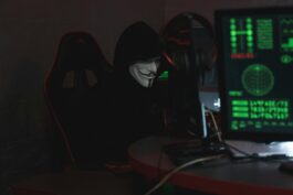 An anonymous hacker wearing a Guy Fawkes mask sits at a computer in a dimly lit room, engaged in cyber activities.