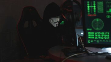 An anonymous hacker wearing a Guy Fawkes mask sits at a computer in a dimly lit room, engaged in cyber activities.