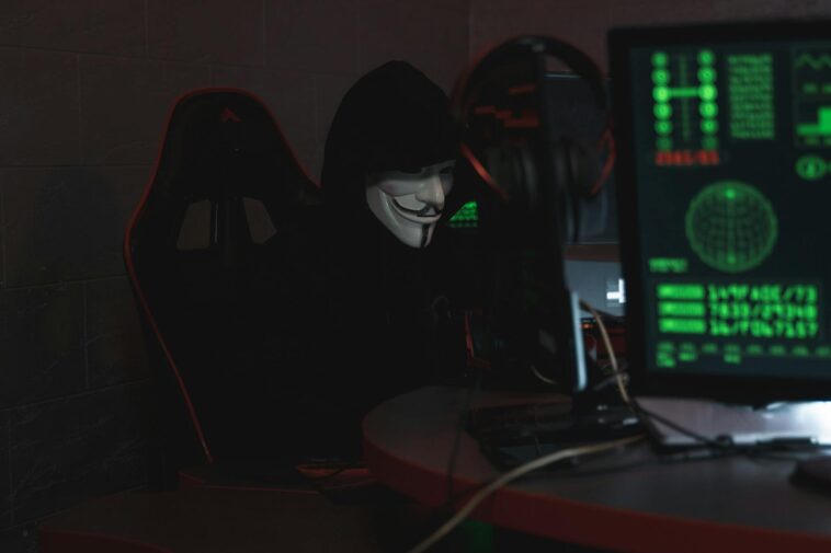 An anonymous hacker wearing a Guy Fawkes mask sits at a computer in a dimly lit room, engaged in cyber activities.