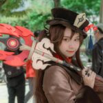 A female cosplayer dressed as Hu Tao from Genshin Impact poses outdoors with a detailed costume and weapon.