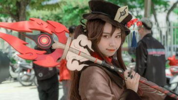 A female cosplayer dressed as Hu Tao from Genshin Impact poses outdoors with a detailed costume and weapon.