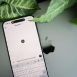 A smartphone shows a ChatGPT interface placed on an Apple laptop in a leafy environment.