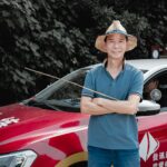 A man with a straw hat stands confidently by a red car, set against a leafy backdrop.