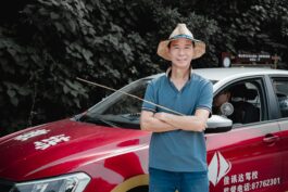 A man with a straw hat stands confidently by a red car, set against a leafy backdrop.