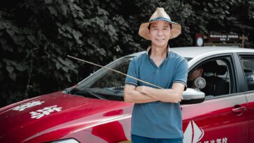 A man with a straw hat stands confidently by a red car, set against a leafy backdrop.