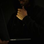 Crop contemplative male hacker in black hoodie watching netbook and touching chin thoughtfully in dark room