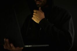 Crop contemplative male hacker in black hoodie watching netbook and touching chin thoughtfully in dark room