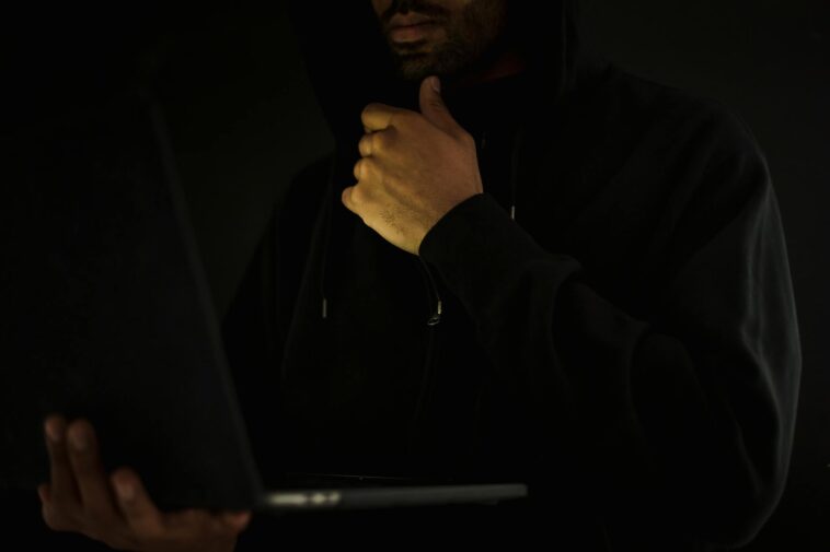 Crop contemplative male hacker in black hoodie watching netbook and touching chin thoughtfully in dark room
