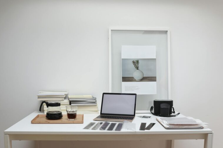 Modern laptop with blank screen arranged with retro filmstrips photo camera and framed picture placed on table near coffee and stack of books