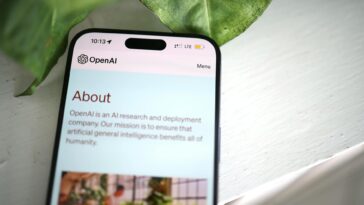Close-up shot of a smartphone screen showing the OpenAI website with greenery in the background.