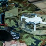 Compact white drone on camouflage gear, surrounded by flashlight and tools.